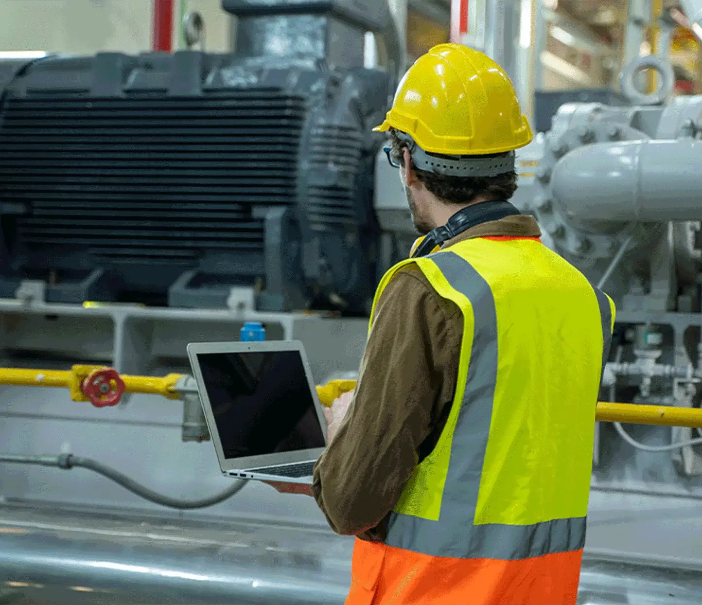 Worker in safety gear inspecting machinery with laptop.