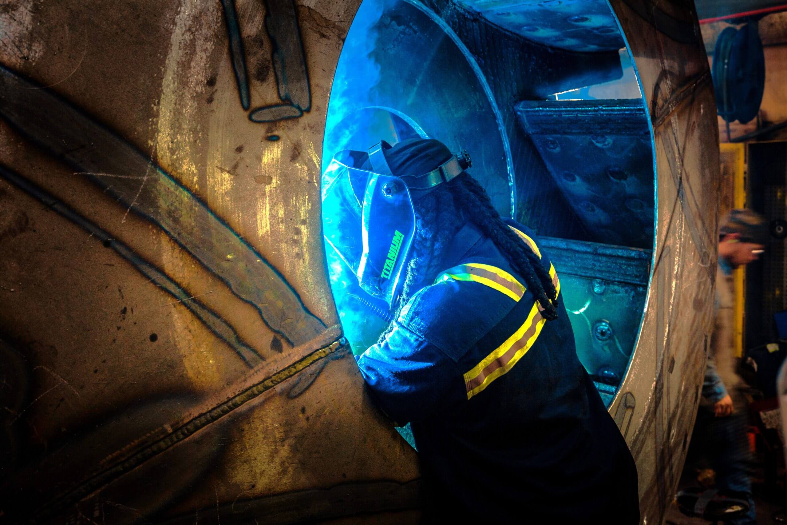 Welder works on large industrial machinery, sparks flying.